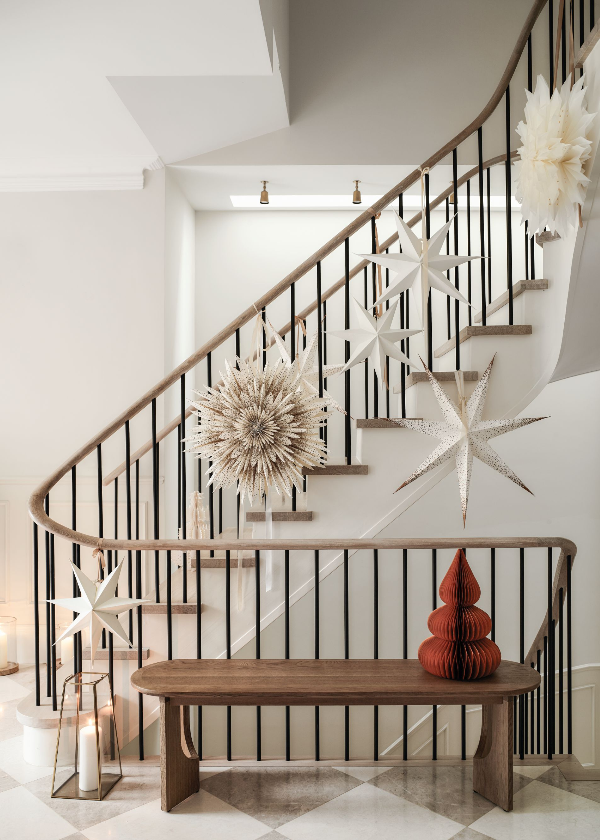 White and cream paper lampshades and stars are attached to a stair rail with a red paper christmas tree decoration sitting on top of a wooden bench at the bottom