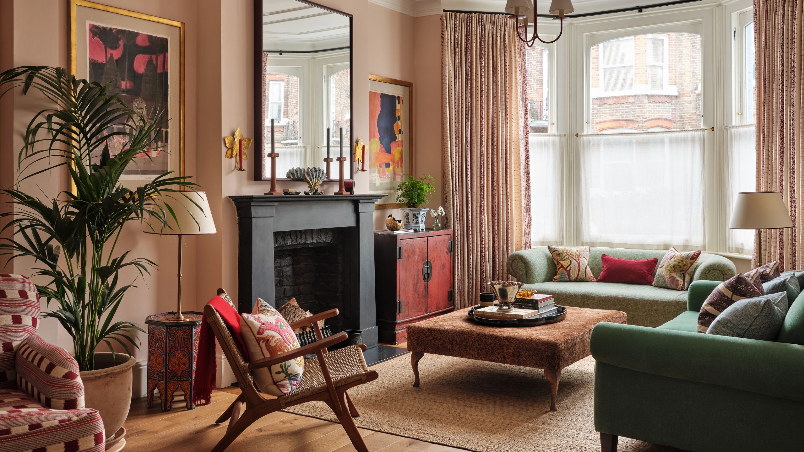 a large warm pink victorian living room with bay window, fireplace and detailed moldings styled with green couches and unique art and vintage furniture