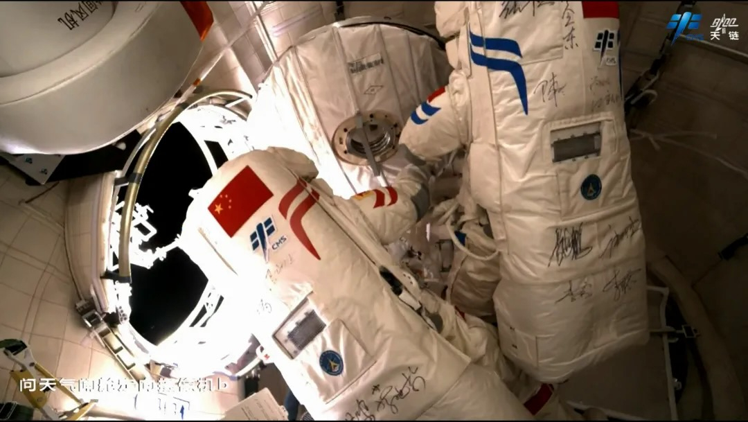 two astronauts in white spacesuits inside a space station, with a circular hatch open and the blackness of space visible outside