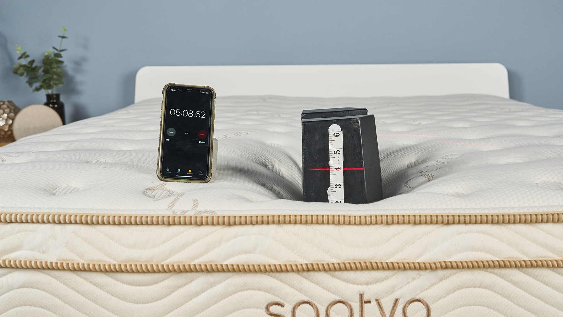 This image shows a close up of our edge support tests on the Saatva Classic Mattress. A 25kg weight is balanced at the foot of the Saatva Classic Mattress. To the left, a phone is balanced displaying a timer at 5 minutes and 8 seconds. A tape measure is attached to the end of the weight and a laser (out of shot) is directed at the tape measure, hitting just below the 4 inch mark. This shows that after five minutes of testing, the weight has only sank 4 inches into the bed.