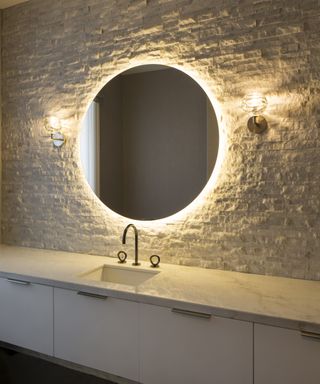 A powder room featuring a large, circular, backlit mirror on a textured stone wall, with black taps
