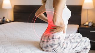 A woman sits on the edge of her mattress aches holding her sore lower back