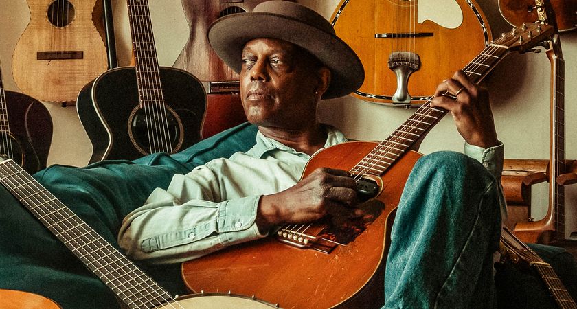 A portrait of Eric Bibb reclining on his sofa, picking on an acoustic guitar, and surrounded by his guitar collection.