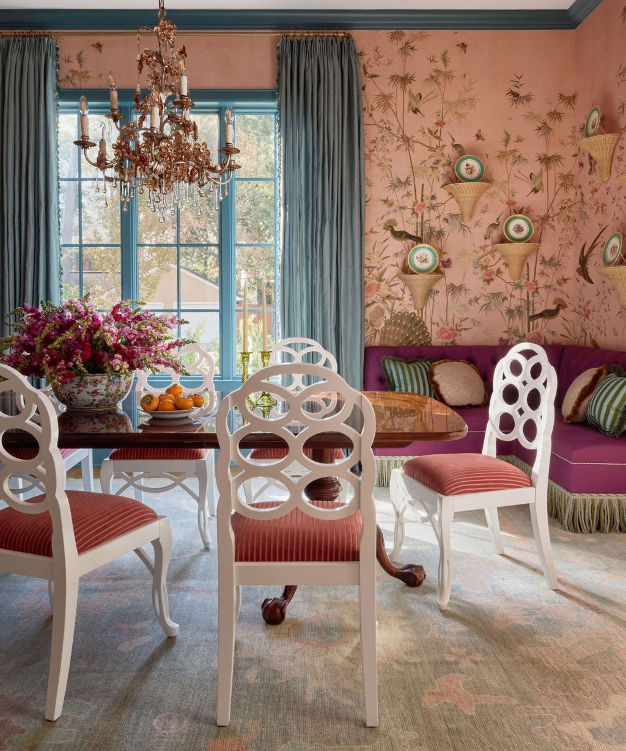Bold dining room with pink bird wallpaper, a vibrant purple banquette, white patterned chairs, and a elaborate chandelier