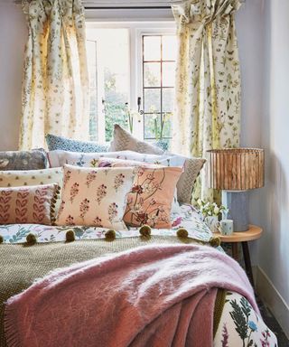 Bed covered with floral pillows and bedcovers and woollen throws beside an open window.