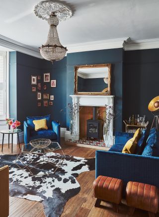 A blue living room with velvet sofa and armchair in the corner with cow print rug and chandelier ceiling light decor