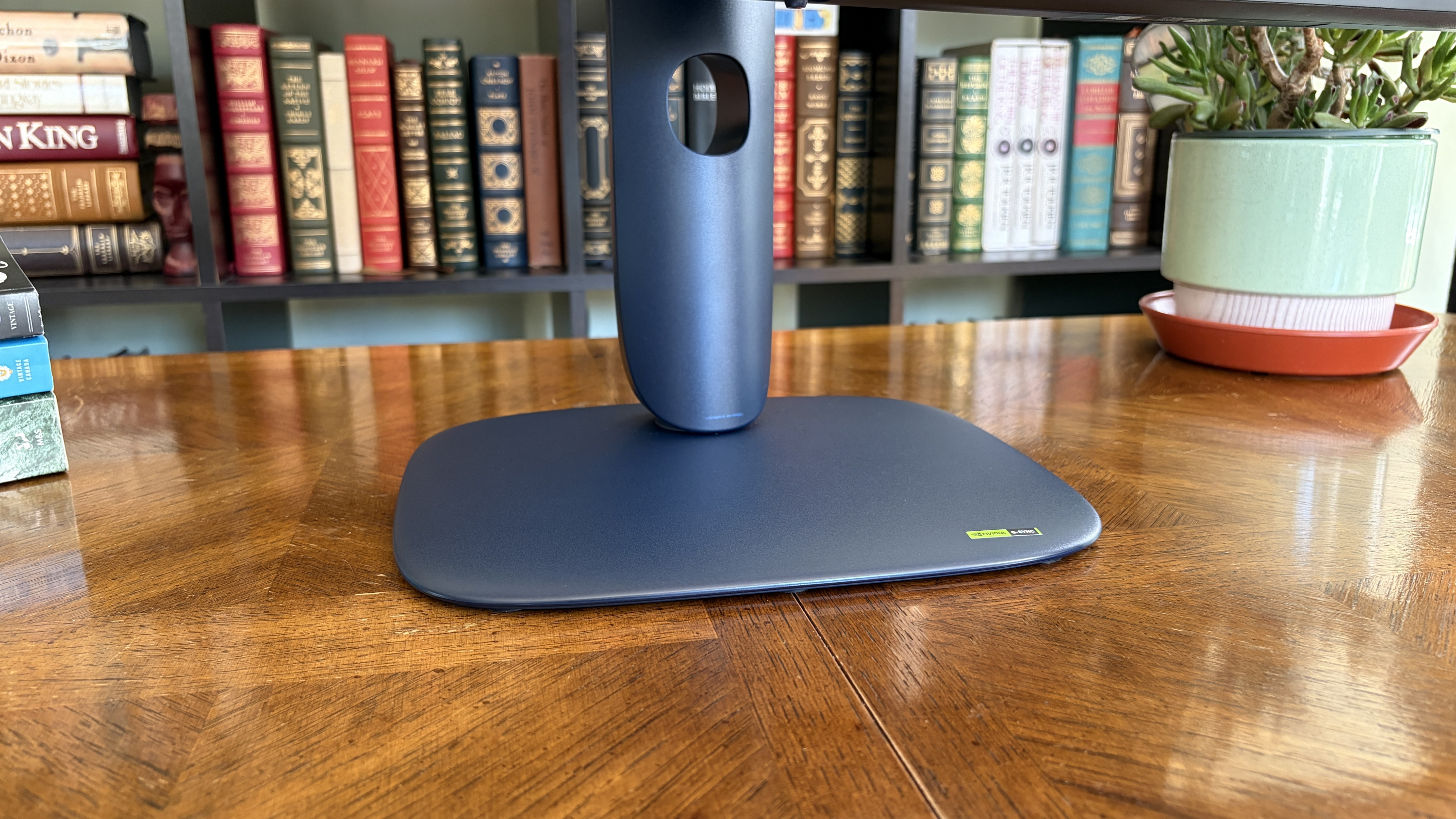 Close-up of a navy blue Alienware AW3426DW monitor stand on a wooden table with bookshelves in the background. A green potted plant is visible to the right.