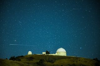 A long white train from a perseid meteor passes over the domed structure of the Lick Observatory in a horizontal orientation against a starry sky.
