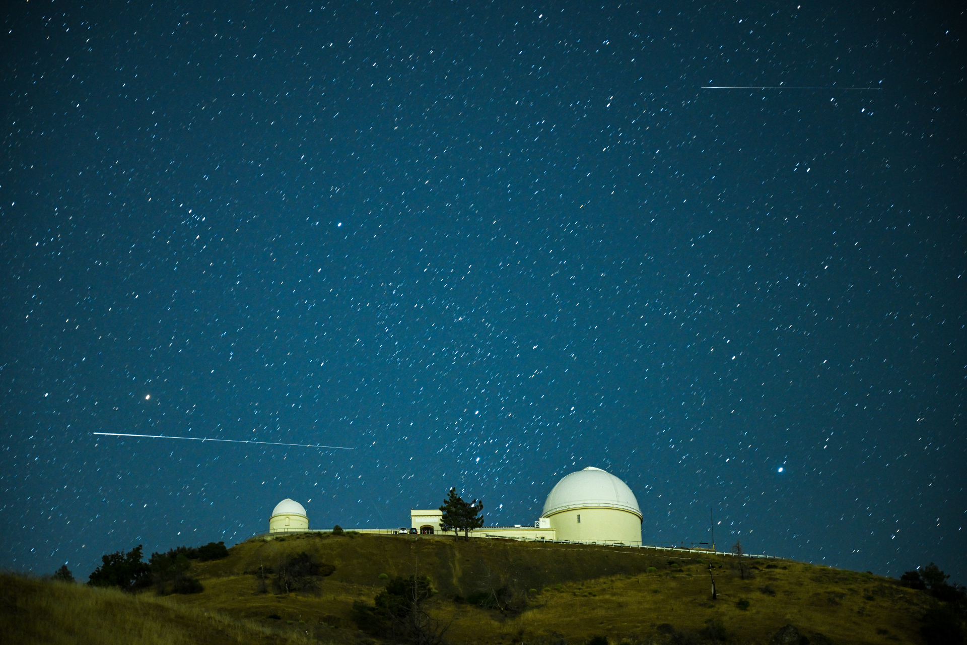ペルセウス座流星の白く長い列が、星空を背景に湖天文台のドーム型構造物の上を水平方向に通過します。
