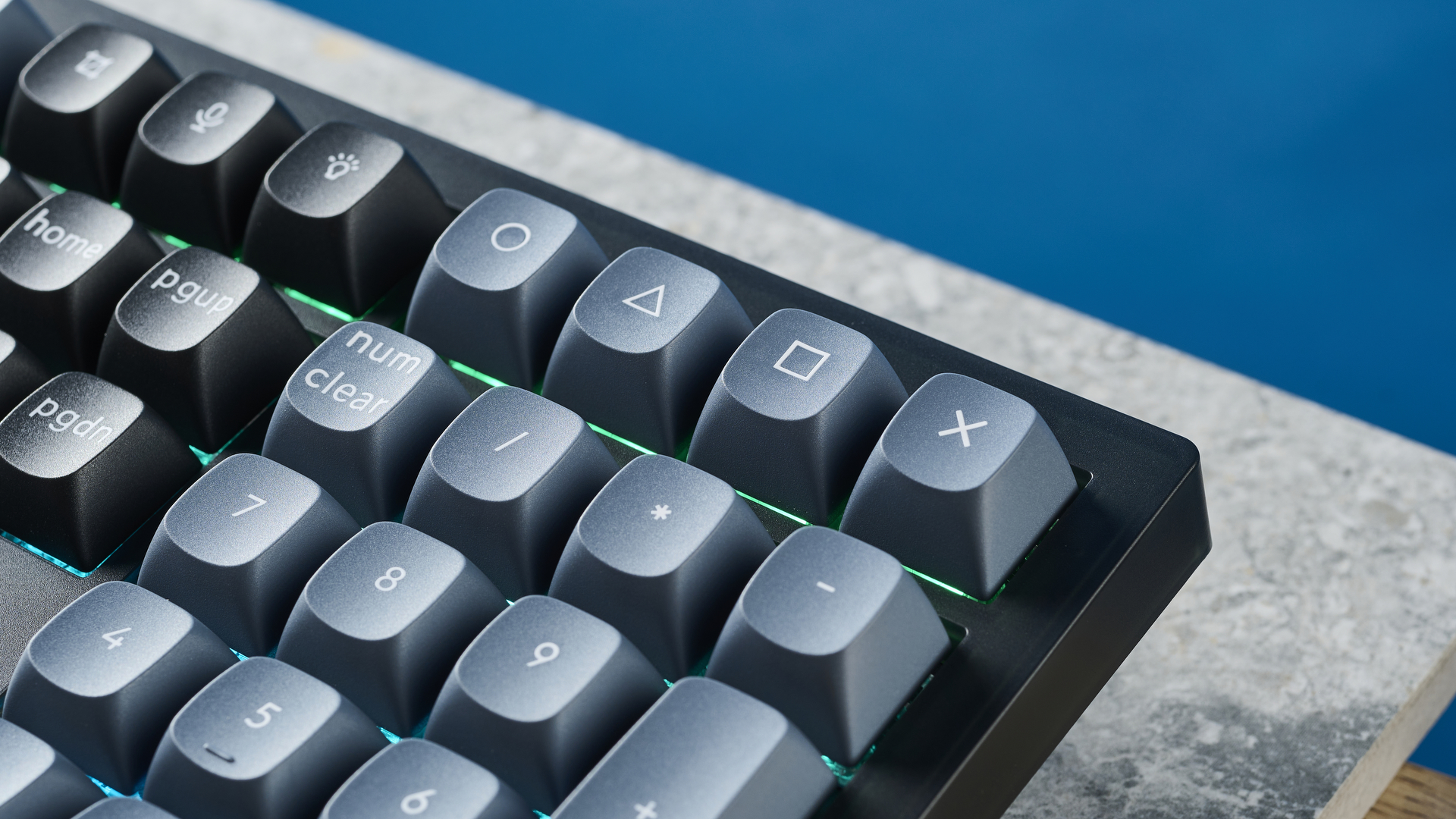 A photograph of the Keychron V6 in black, with gray , black and orange keycaps. The keyboard is positioned on a stone slate, with a blue wall in the background.