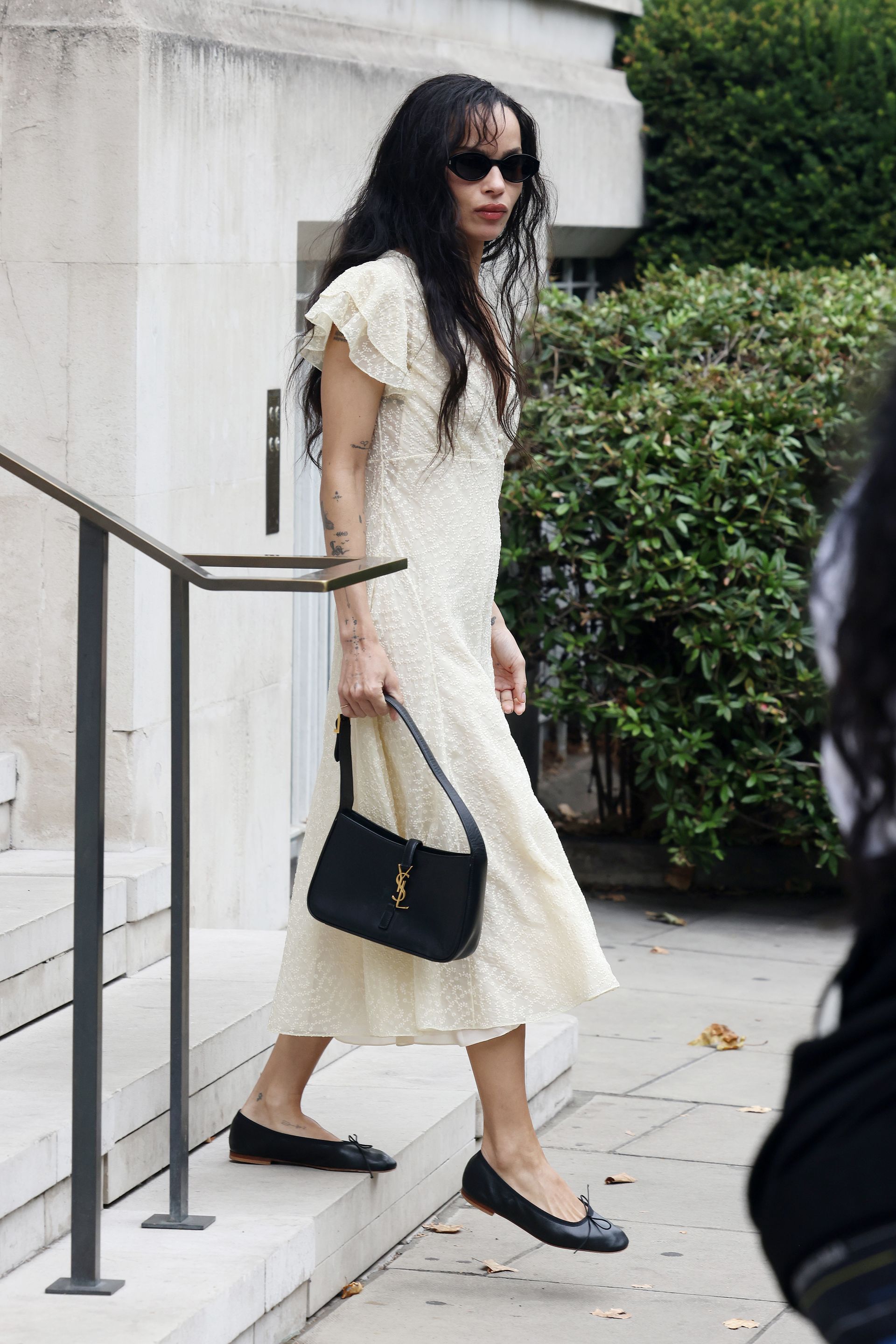 Zoe Kravitz walks down steps outside wearing a butter yellow dress with black ballet flats and a YSL handbag.