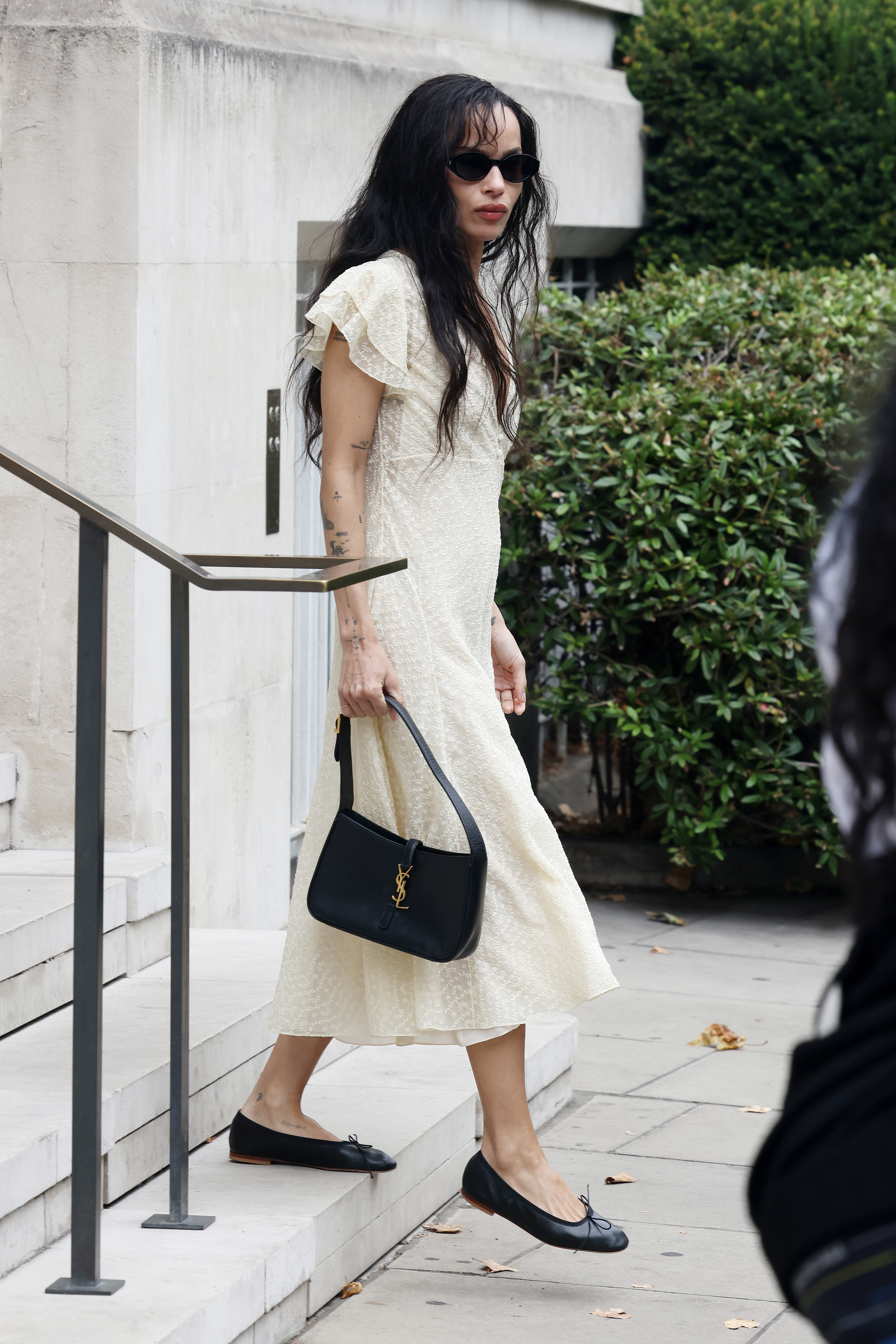 Zoe Kravitz walks down steps outside wearing a butter yellow dress with black ballet flats and a YSL handbag.