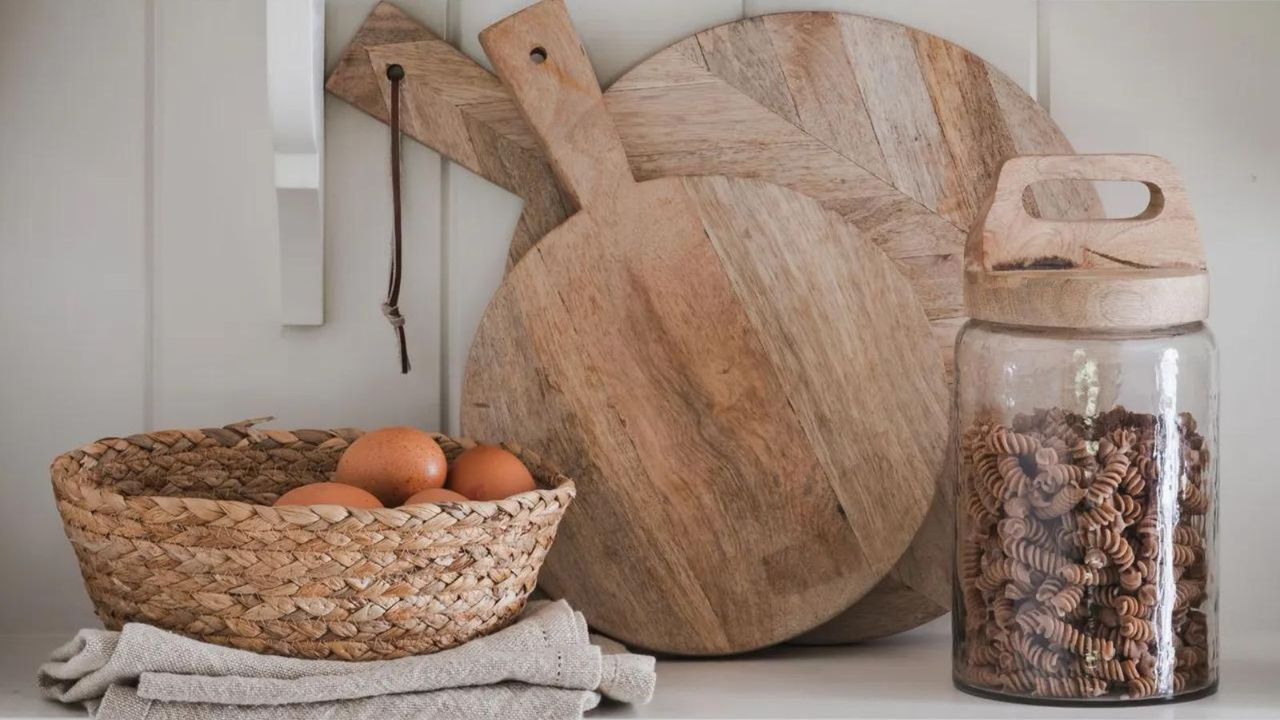 Wooden cutting boards stacked on a kitchen shelf with a basket of eggs and a jar of dried pasta