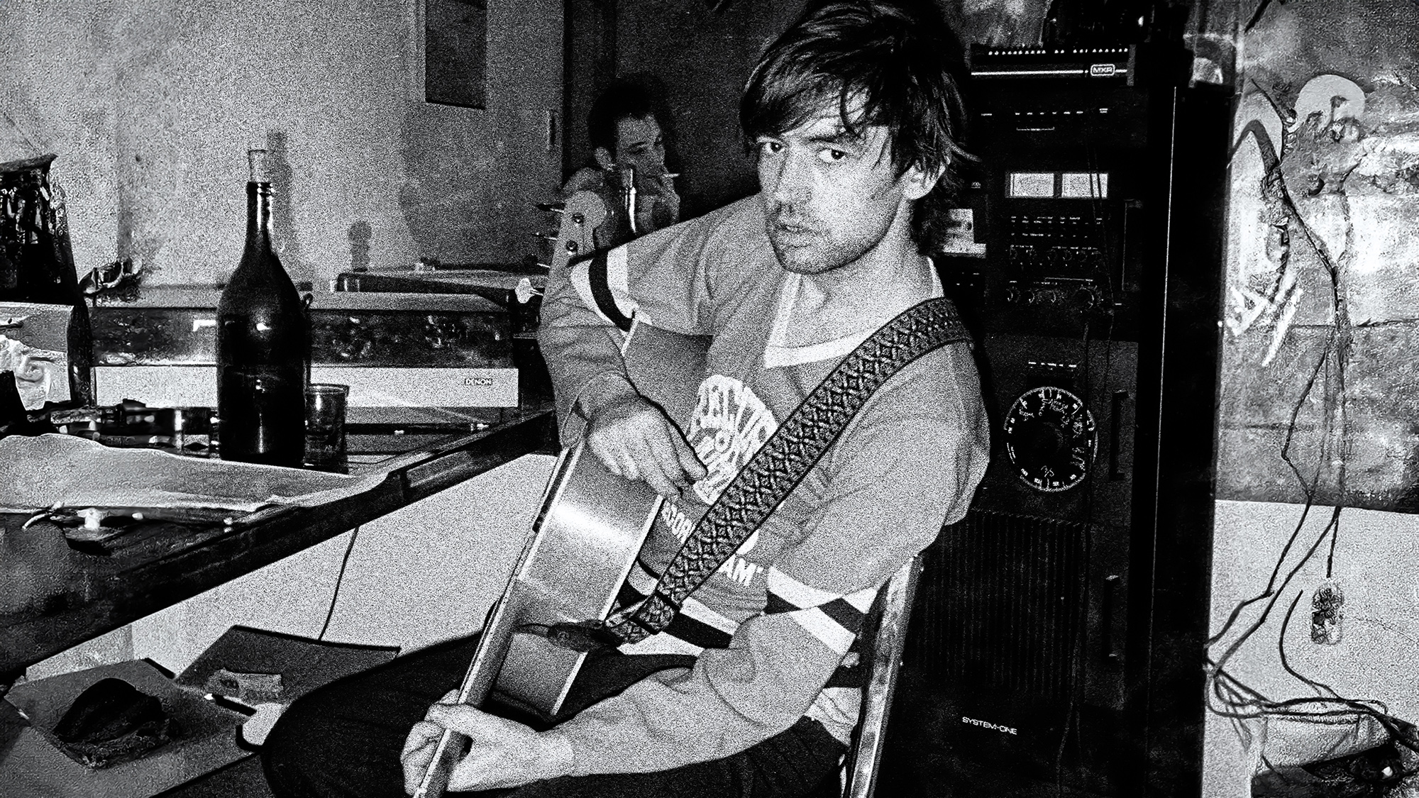 Portrait of British Rock musician Chris Spedding, an acoustic guitar in his lap, as he sits at home, New York, New York, 1982. Singer Robert Gordon, with whom Spedding performed, is in the background.