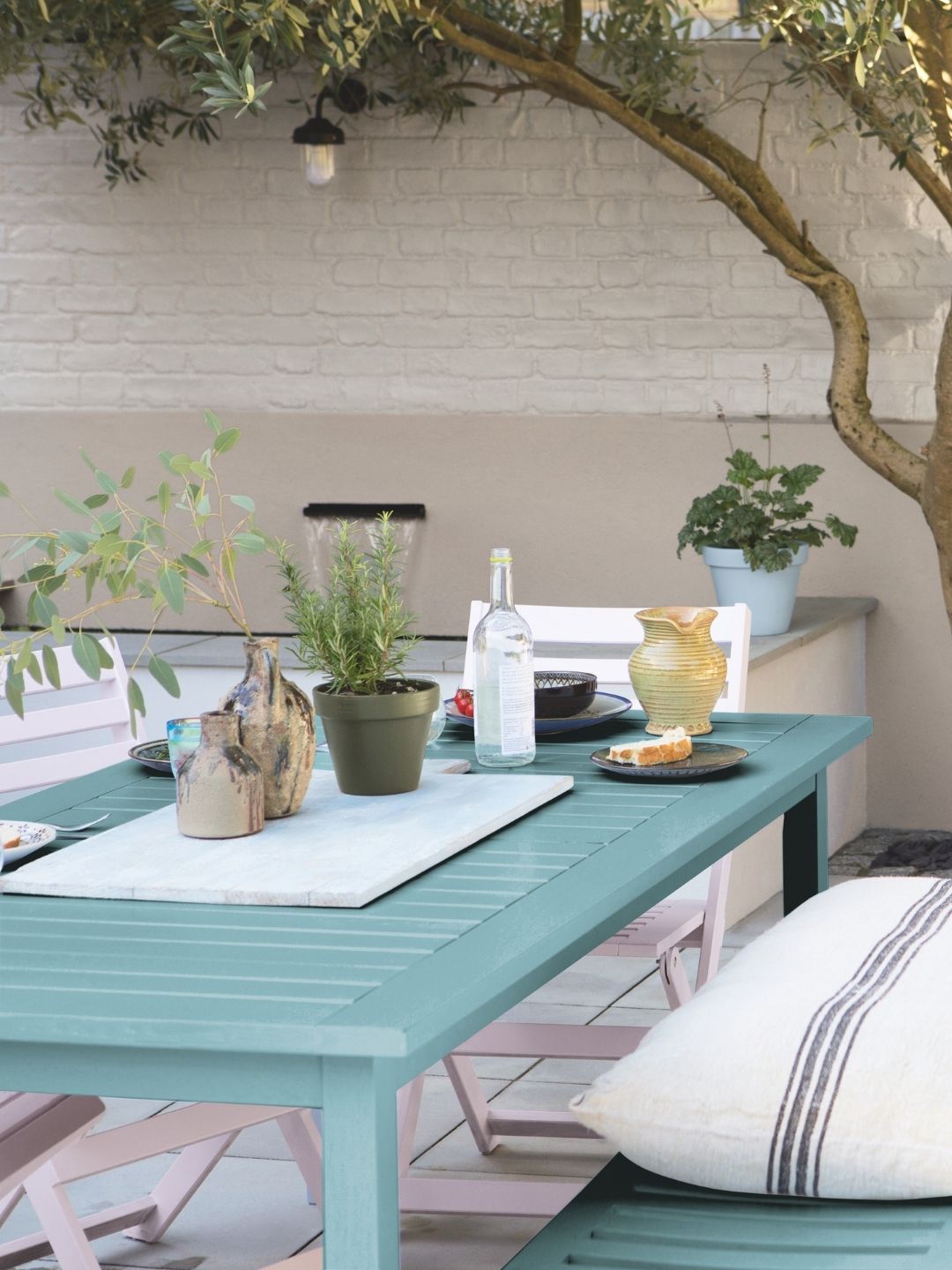 Image of a teal outdoor table with dining supplies on it and a few light pink chairs around it. The patio is painted in a beige color.