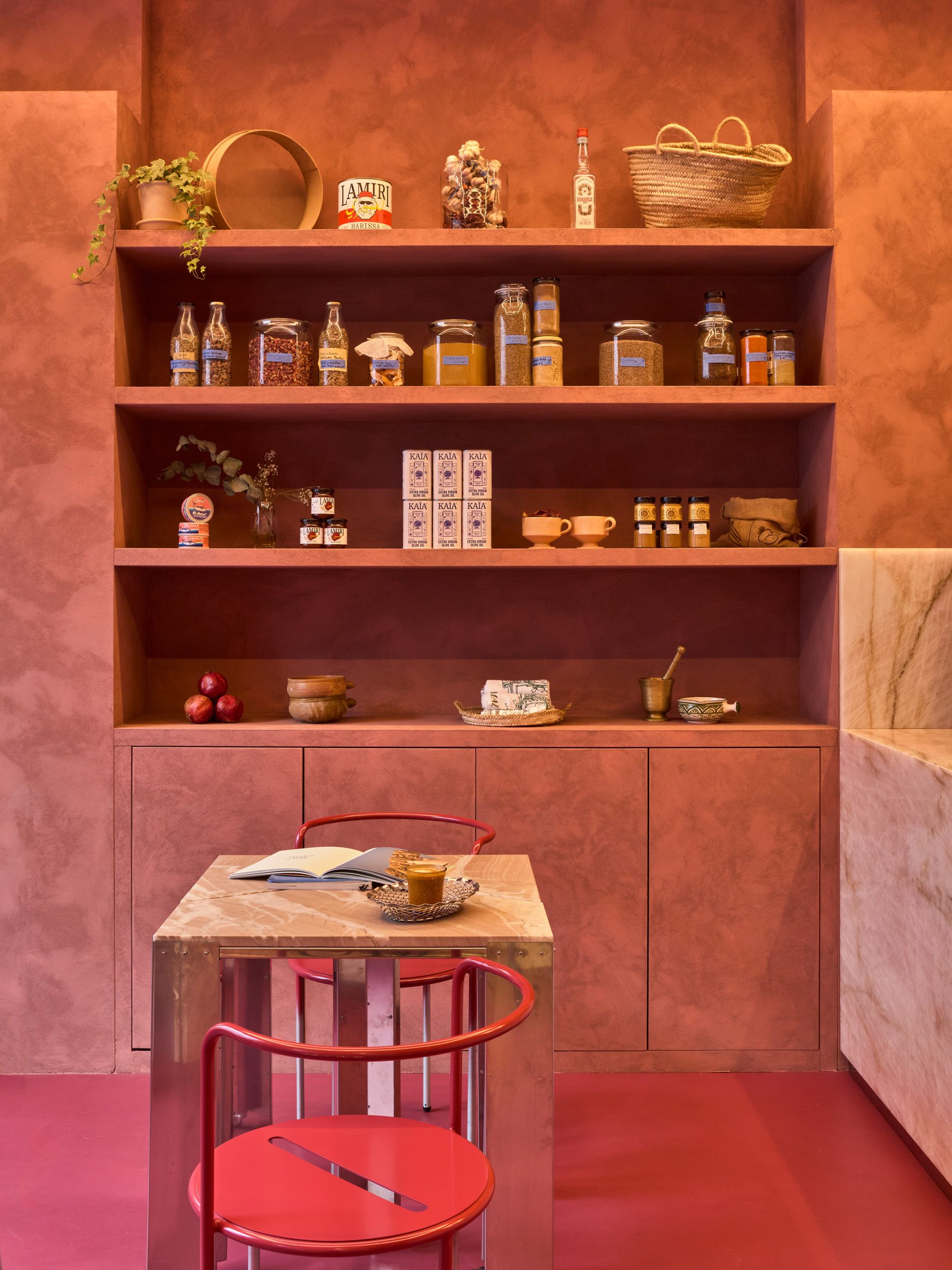 A pink-painted cafe with bright pink and chrome chairs, marble-topped sculptural tables with chrome legs, and lots of deli products and artworks styled on its shelves.