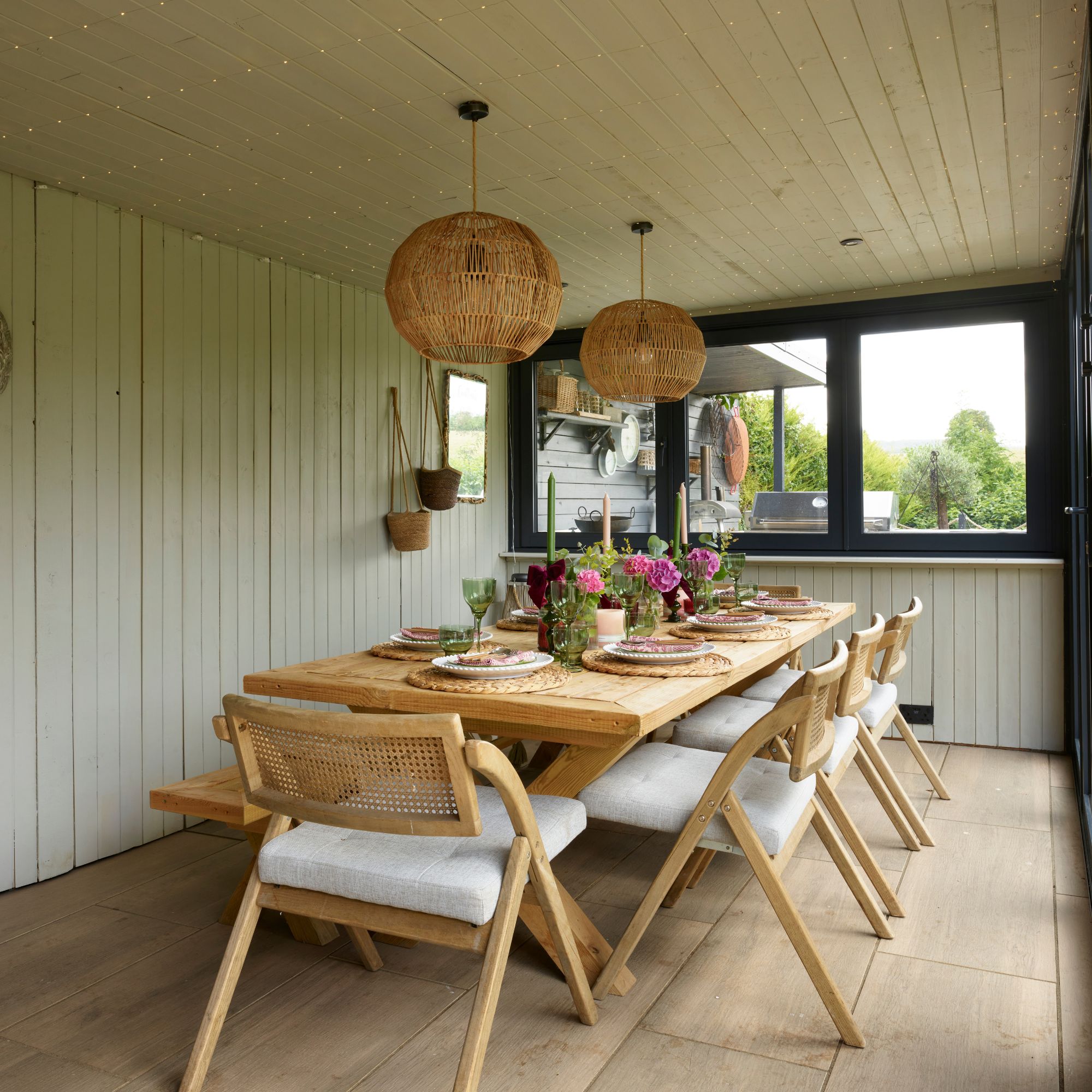 Garden room with green panelled walls and wooden dining table with cane chairs. Rattan lighting with black windows.