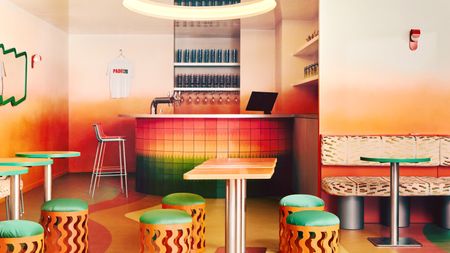 A bar with gradient floors, walls, and bar tiles ranging from red to yellow and green features quirky furniture like wavy stools, chrome-clad tables with a colorful top, curious mirrors, and animalier-style banquettes in orange, cream, and black.