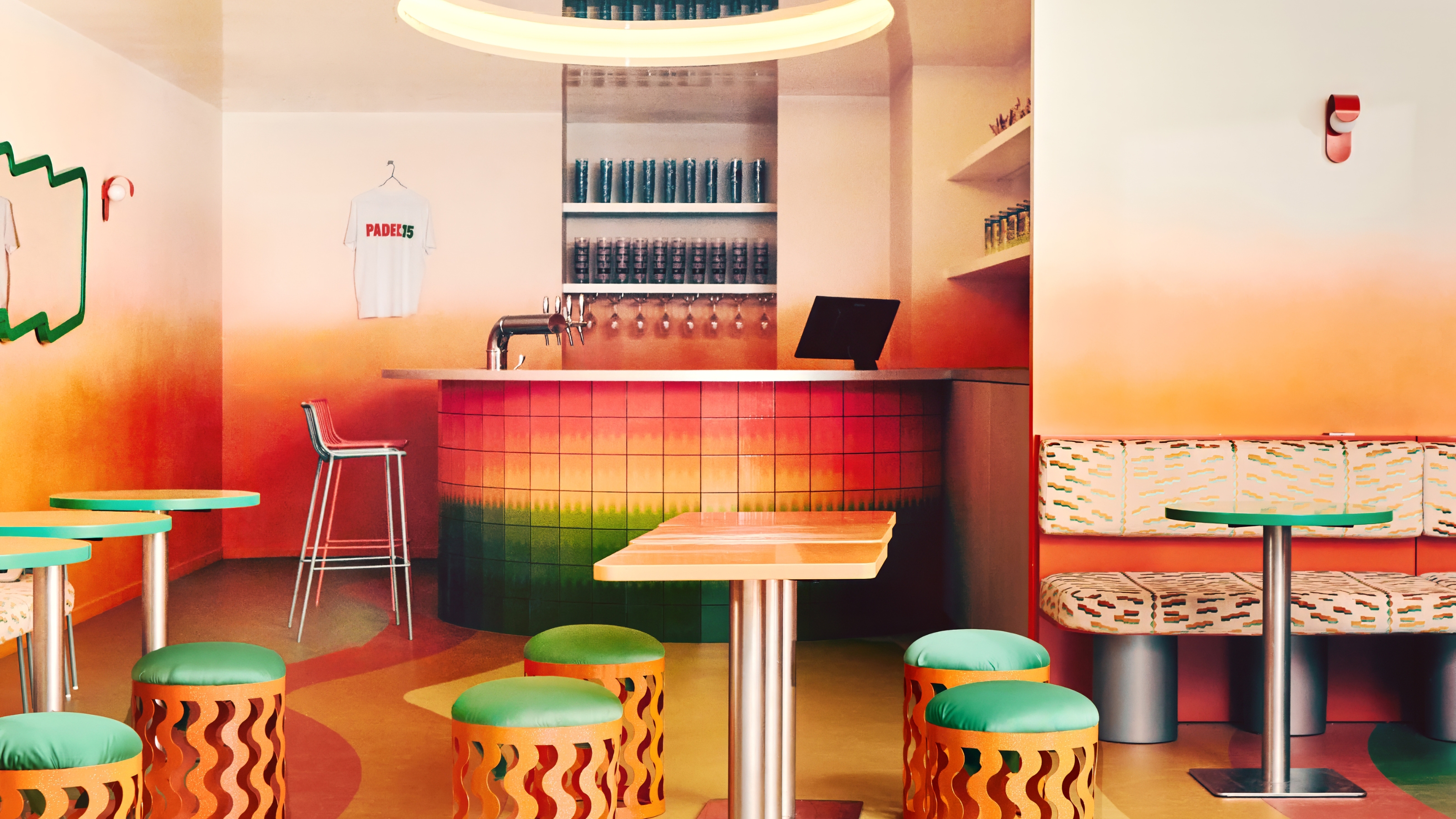 A bar with gradient floors, walls, and bar tiles ranging from red to yellow and green features quirky furniture like wavy stools, chrome-clad tables with a colorful top, curious mirrors, and animalier-style banquettes in orange, cream, and black.