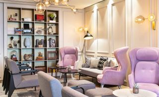 A living area in the Hilton London Bankside hotel. Light gray and pink chairs, with black and white marble coffee tables, take up the entire room. There is a shelf on the wall to the left with various decorations on it.