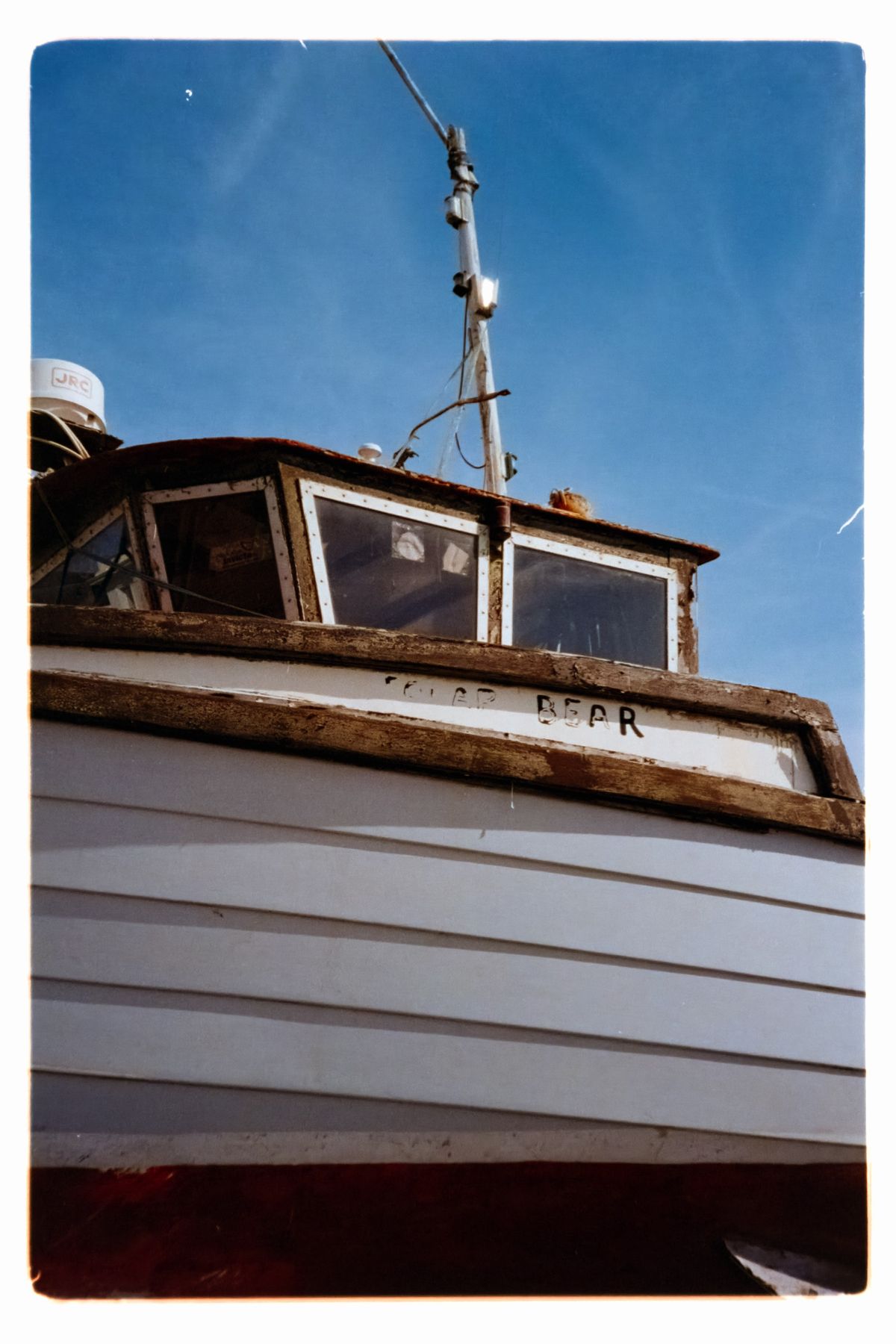 Scanned film close-up of the upper section of a weathered fishing boat, including its cabin, mast, and navigation equipment against a blue sky.