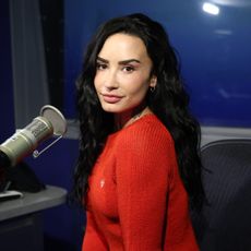 a white woman with dark hair wearing a bright red sweater and posing in front of a microphone in a studio