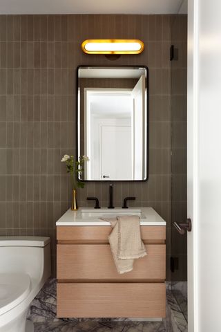 An earthy wall tile in a modern bathroom with wooden vanity unit and warm lighting above the mirror