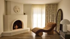 A white living room with a a white arched fireplace, a brown fainting sofa, a white mushroom lamp, and a bobbin-framed arched mirror