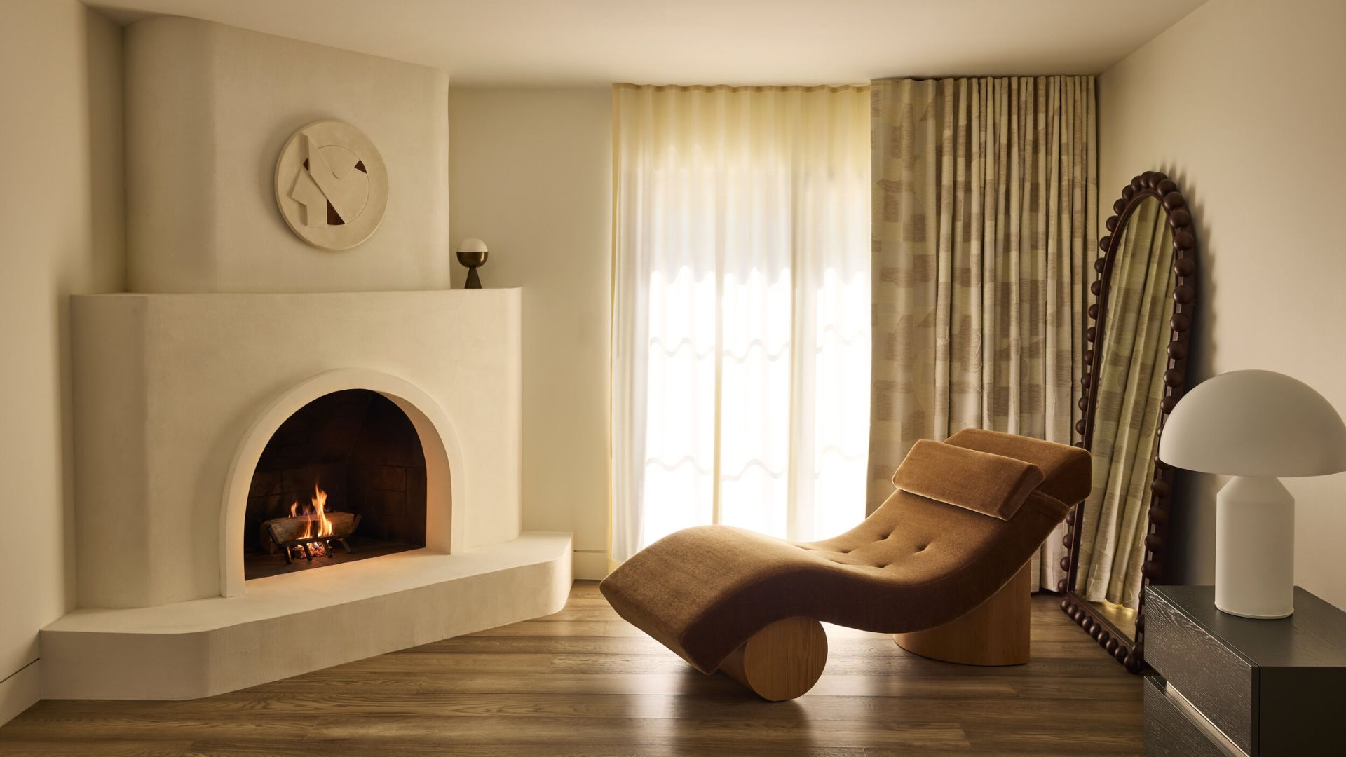 A white living room with a a white arched fireplace, a brown fainting sofa, a white mushroom lamp, and a bobbin-framed arched mirror