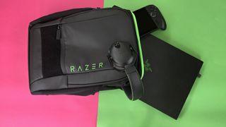 A black Razer backpack, Razer headphones, a black laptop, and a handheld gaming device are placed on a split pink and green background.