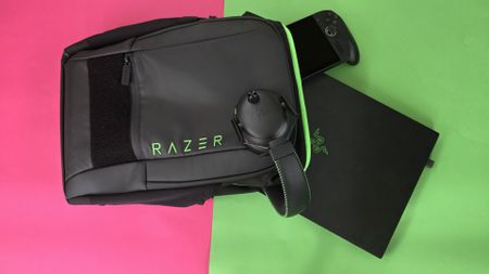 A black Razer backpack, Razer headphones, a black laptop, and a handheld gaming device are placed on a split pink and green background.