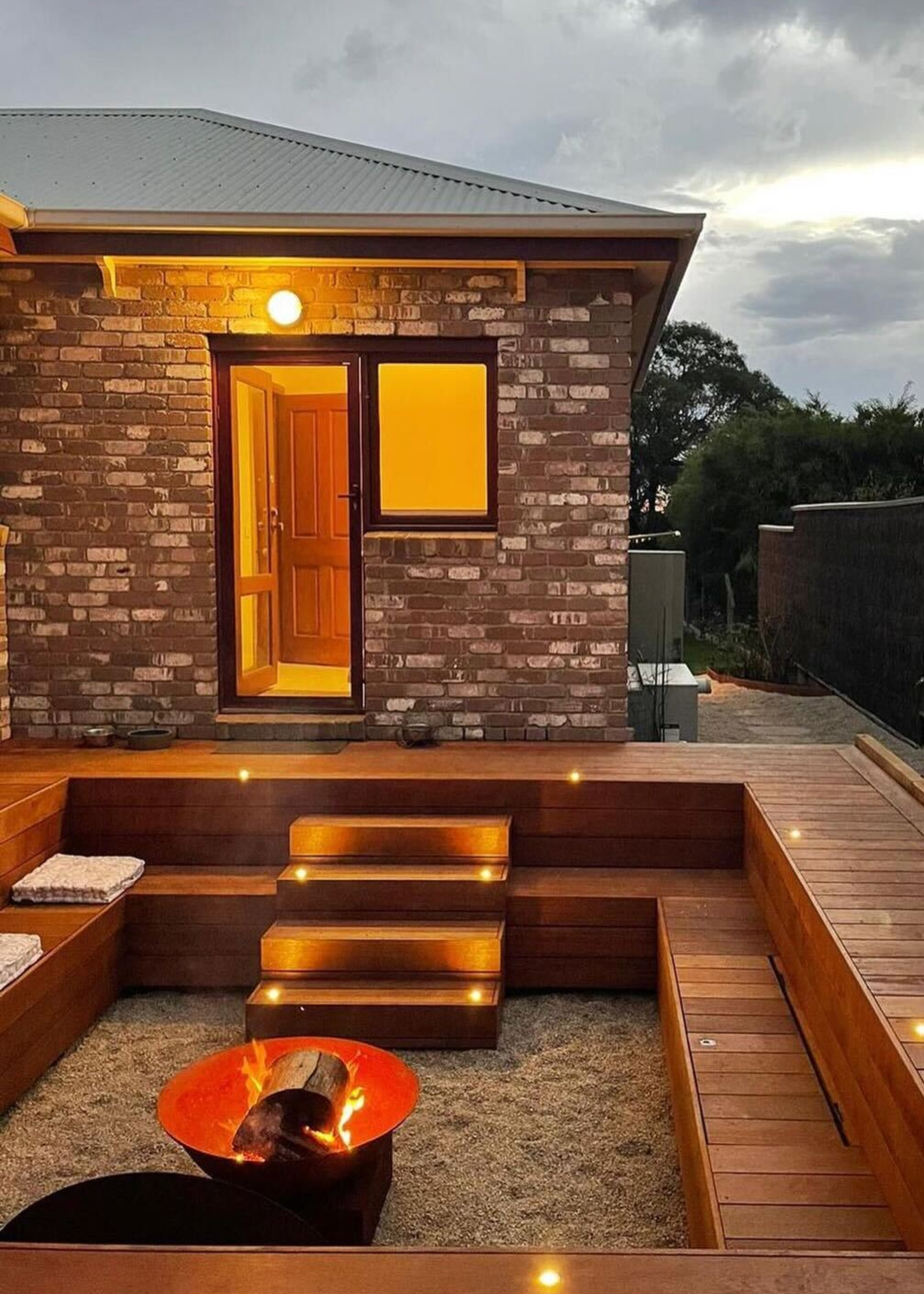 A sunken seating decking are with steps that lead to a building. In the middle of the sunken seating is a firepit.