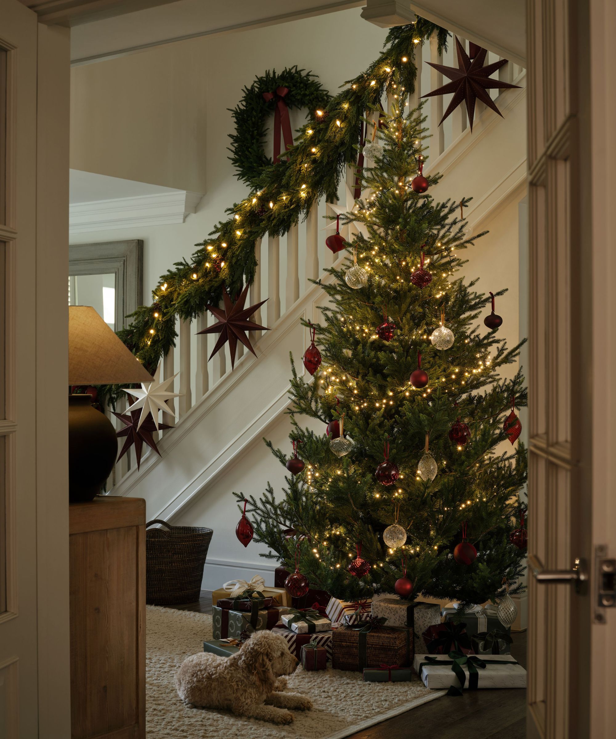 A Christmas tree decorated with red and white baubles in a cream entryway, beside set of stairs with a Christmas garland. A small poodle dog lays next to presents beneath the tree.