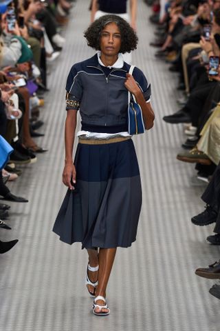 Model wears a pleated skirt on the Miu Miu runway with white flats, a zip-up short-sleeve jacket, and shoulder bag.