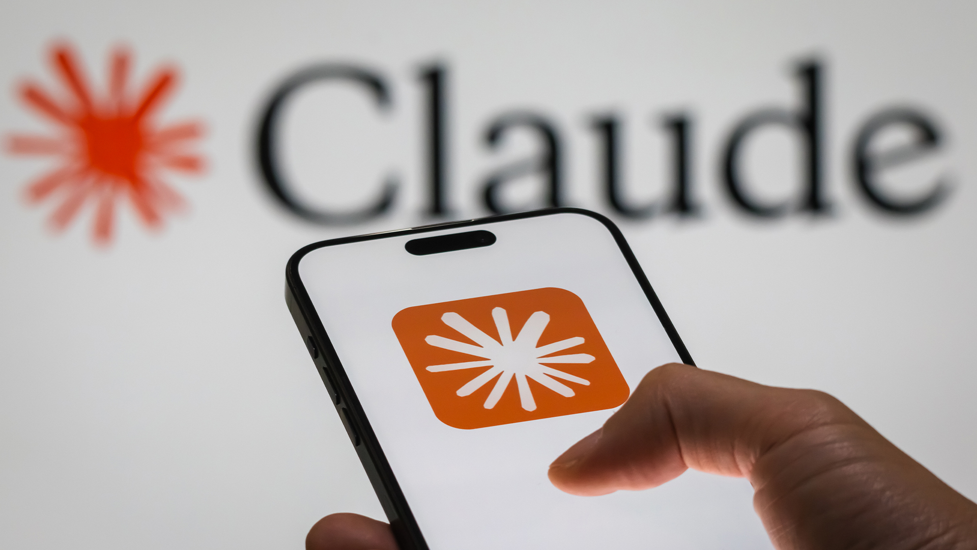 Anthropic Claude logo and branding pictured on a smartphone screen held in hand, with separate branding pictured in background.