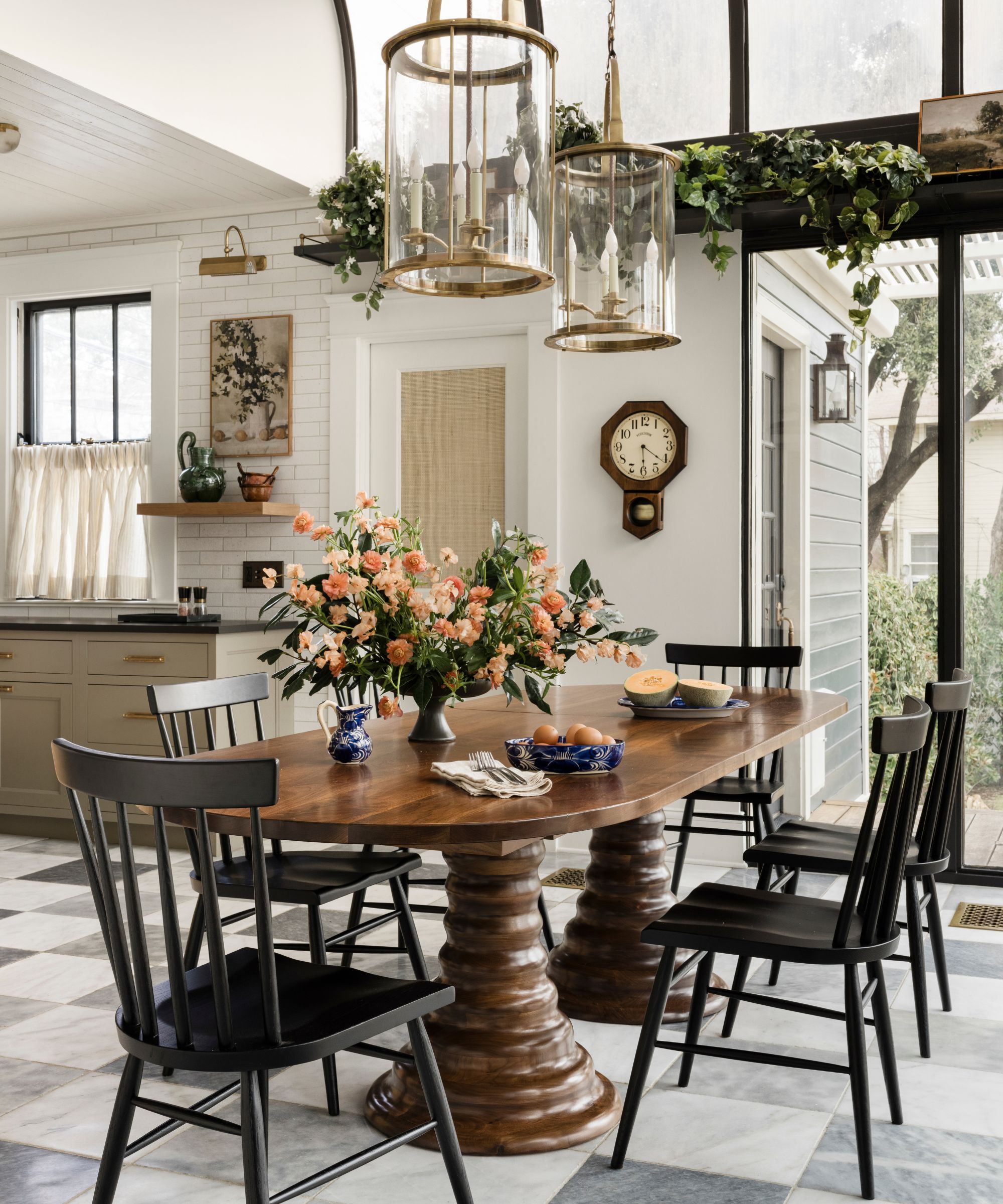 a conservatory off a kitchen with checkerboard stone floors, a large wooden table with two pedestal legs, black wooden chairs and two brass lanterns overhead