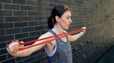 woman in a grey vest standing sideways to the camera against a brick wall stretching a red long loop resistance band across her chest