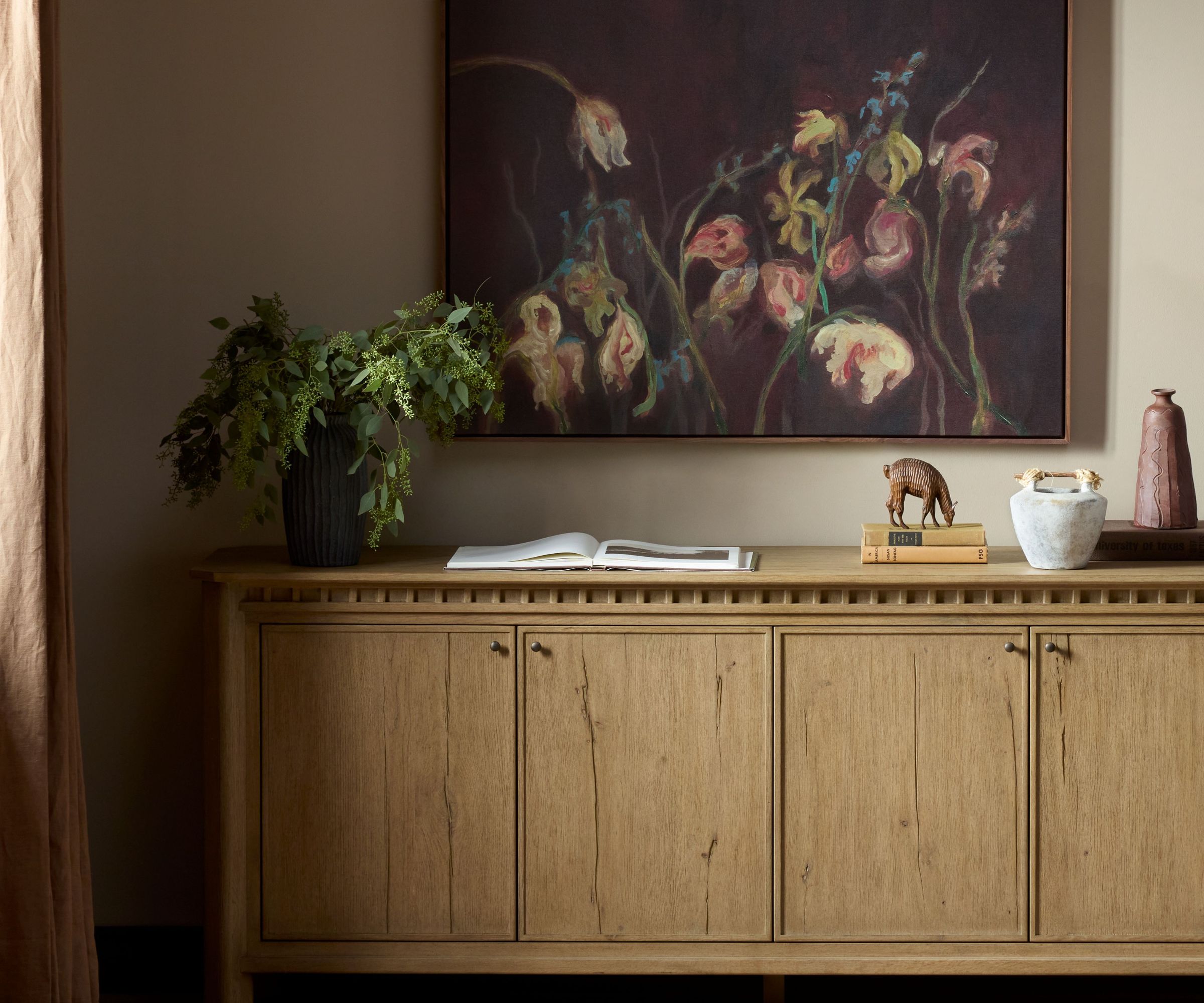 Rustic wooden console unit adorned with a vase of foliage, an open book, a stack of books with an animal ornament, and a rustic bowl beside it. A large painting of flowers sits on the wall above.