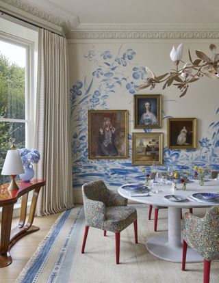 Image of a white dining room that has a blue and white mural-style wallpaper of a pond scene. There is a small gallery wall (four frames) of portrait paintings. The dining table is a modern white table and the dining chairs have a floral-printed upholstery with red legs.