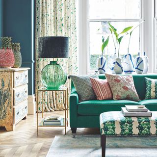 Blue living room with green sofa, a patterned footstool coffee table, and bright curtains in the background