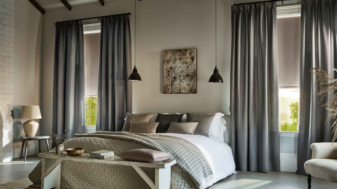 A bedroom with tall ceilings and long drapes, a roller blind beneath. 