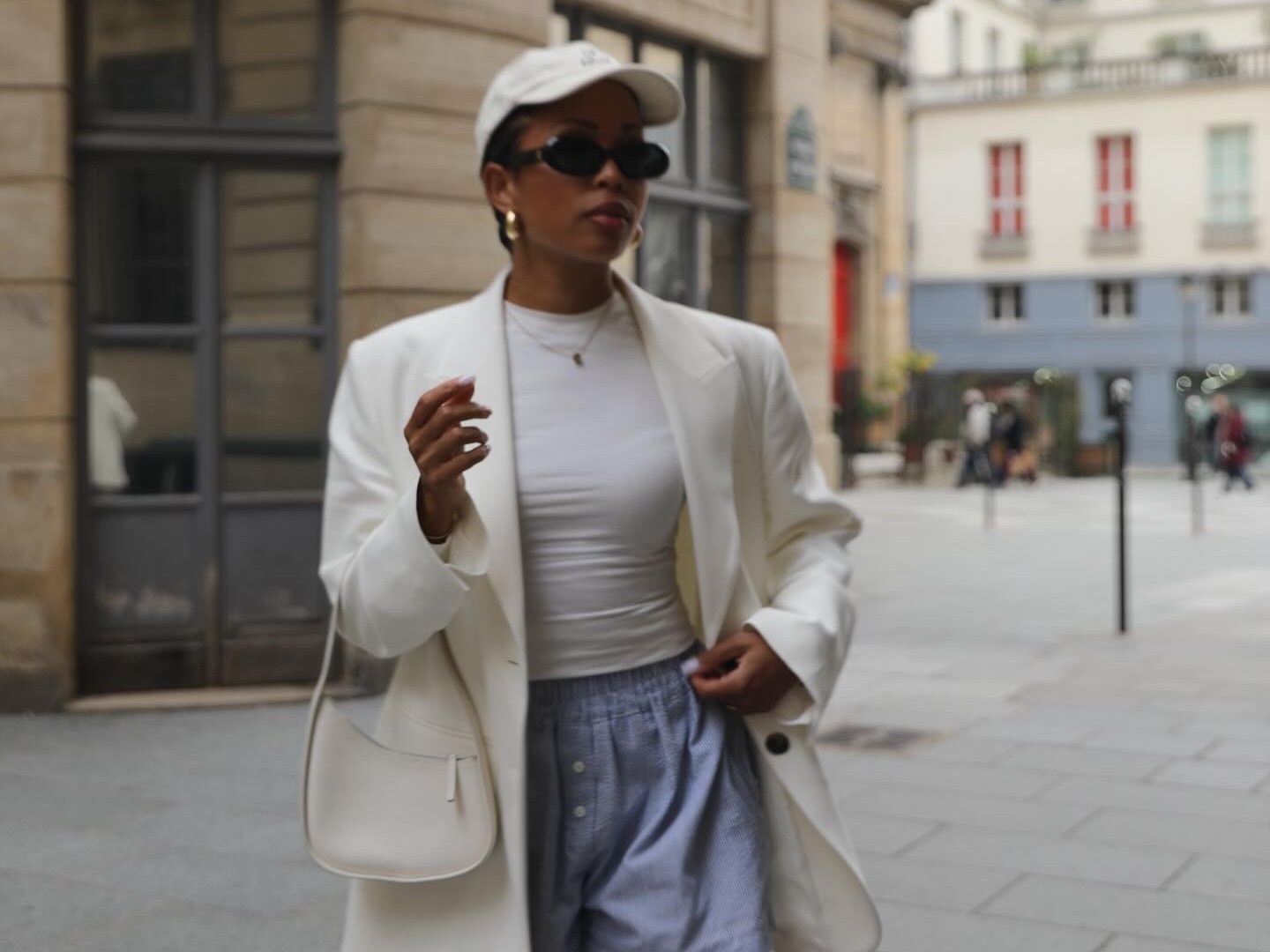 Paris woman wears blue boxer shorts, a fitted white t-shirt, a blazer, and a baseball cap with '90s style black sunglasses and gold hoop earrings.