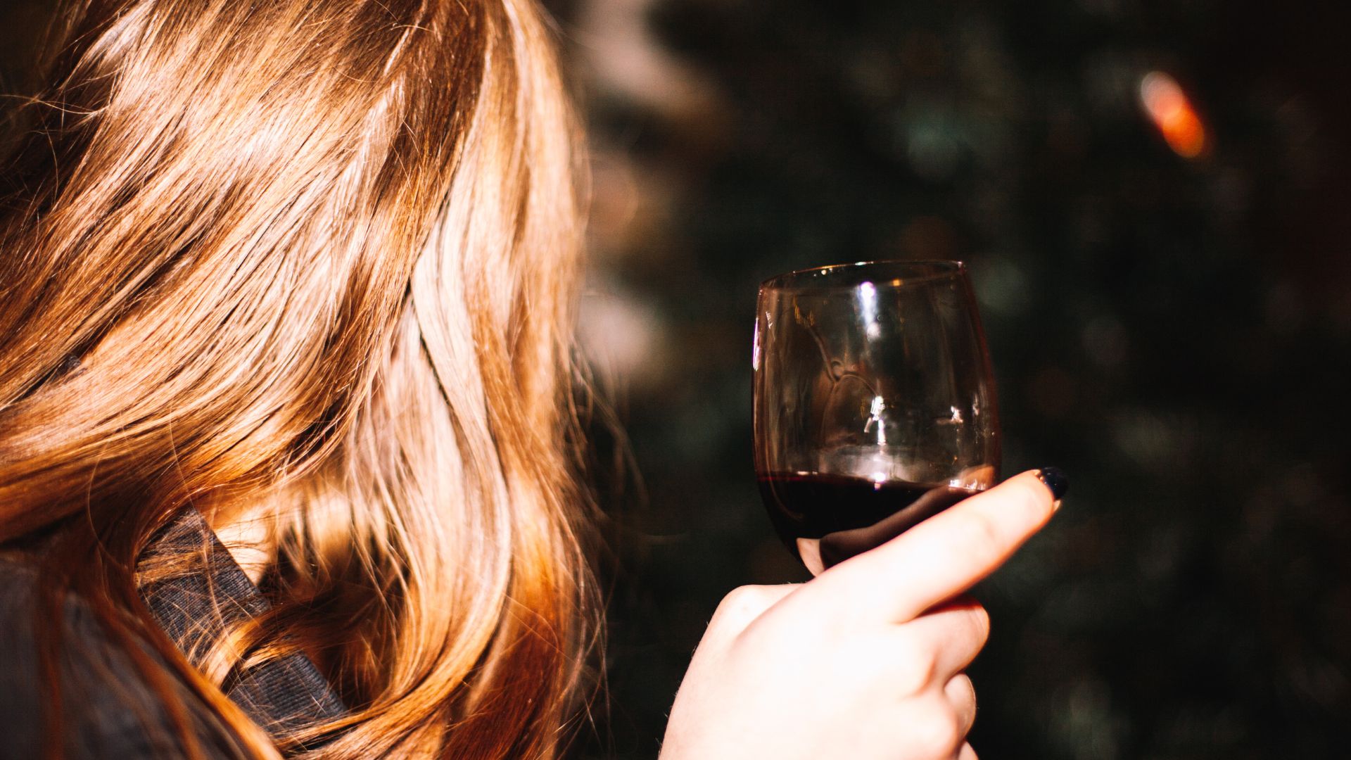 A woman holds a glass of wine at night, she is facing away from the camera.