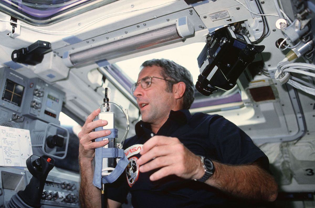 a man in a dark shirt holds what looks like a cordless phone inside the cockpit of a spacecraft