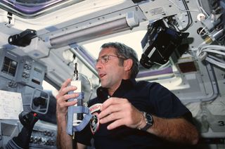 a man in a dark shirt holds what looks like a cordless phone inside the cockpit of a spacecraft