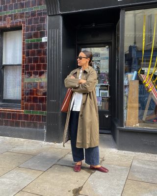 Influencer wears a green trench coat, mules, blue jeans, a white button-down shirt, and a brown shoulder bag.