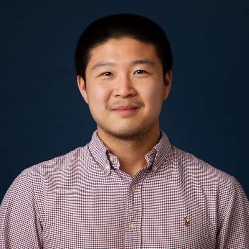 Image of a man in a red and blue plaid shirt standing in front of a dark blue background.