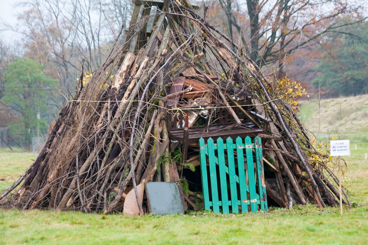 A large bonfire structure made of branches, logs and wooden debris stands on a grassy field, with a small green fence at the front and a sign nearby reading &lsquo;Please no dumping of rubbish&rsquo;.