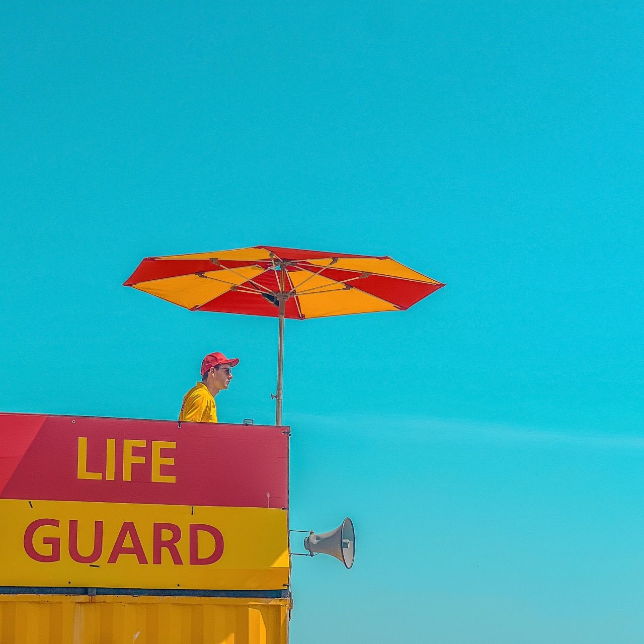 Highly saturated image of a lifeguard in red and yellow under a matching umbrella and above a matching 'Life guard' sign, framed against clear blue sky