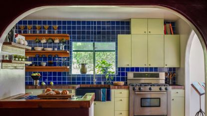 An arched ceiling leading into a kitchen with sage green cabinets, a stainless steel stove and oven, striking cobalt blue tiled backsplash, floating wood shelves adorned with glassware, and a wooden counter